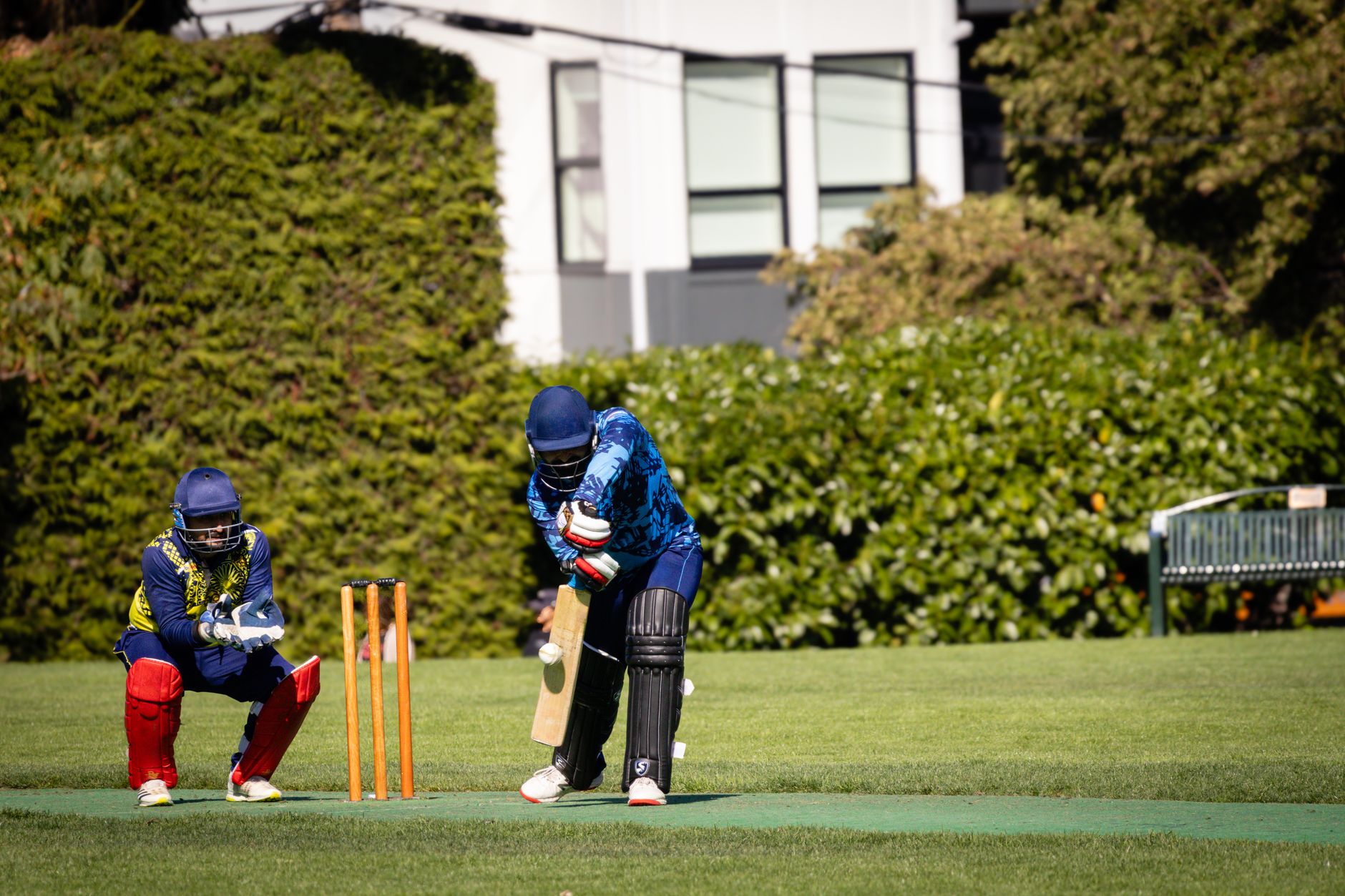 Cricketer batting during an outdoor match with a wicketkeeper behind on a sunny day.
