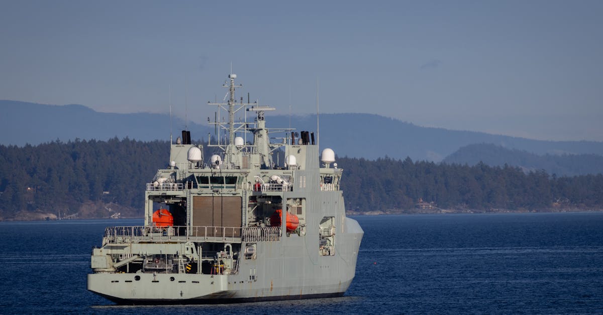 A military warship navigates through open waters with a coastline backdrop on a clear day.