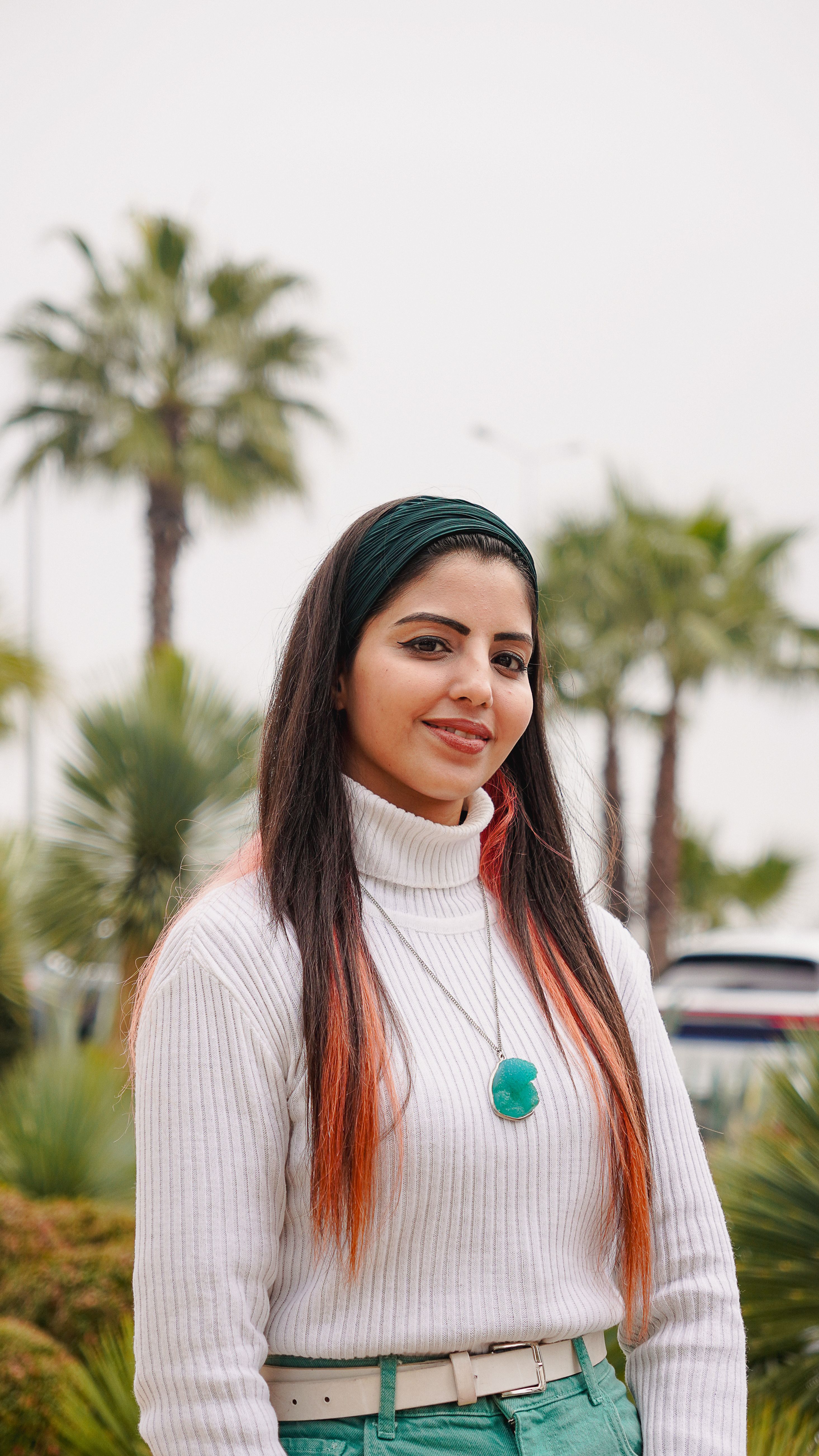 Portrait of a young woman with long hair and a turtleneck sweater, standing outdoors with palm trees.