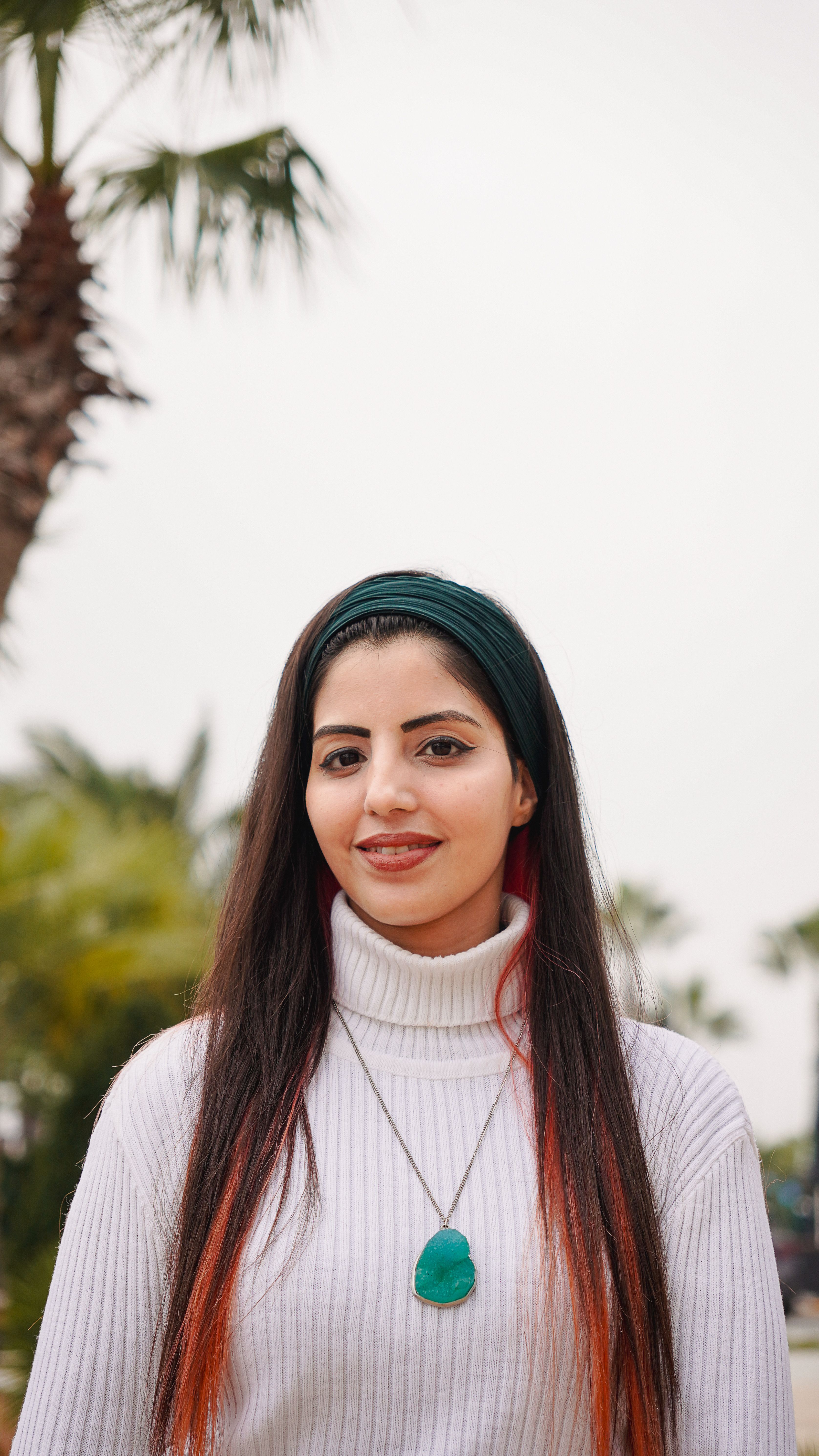 Portrait of a young woman in a white sweater standing outdoors with palm trees in the background.