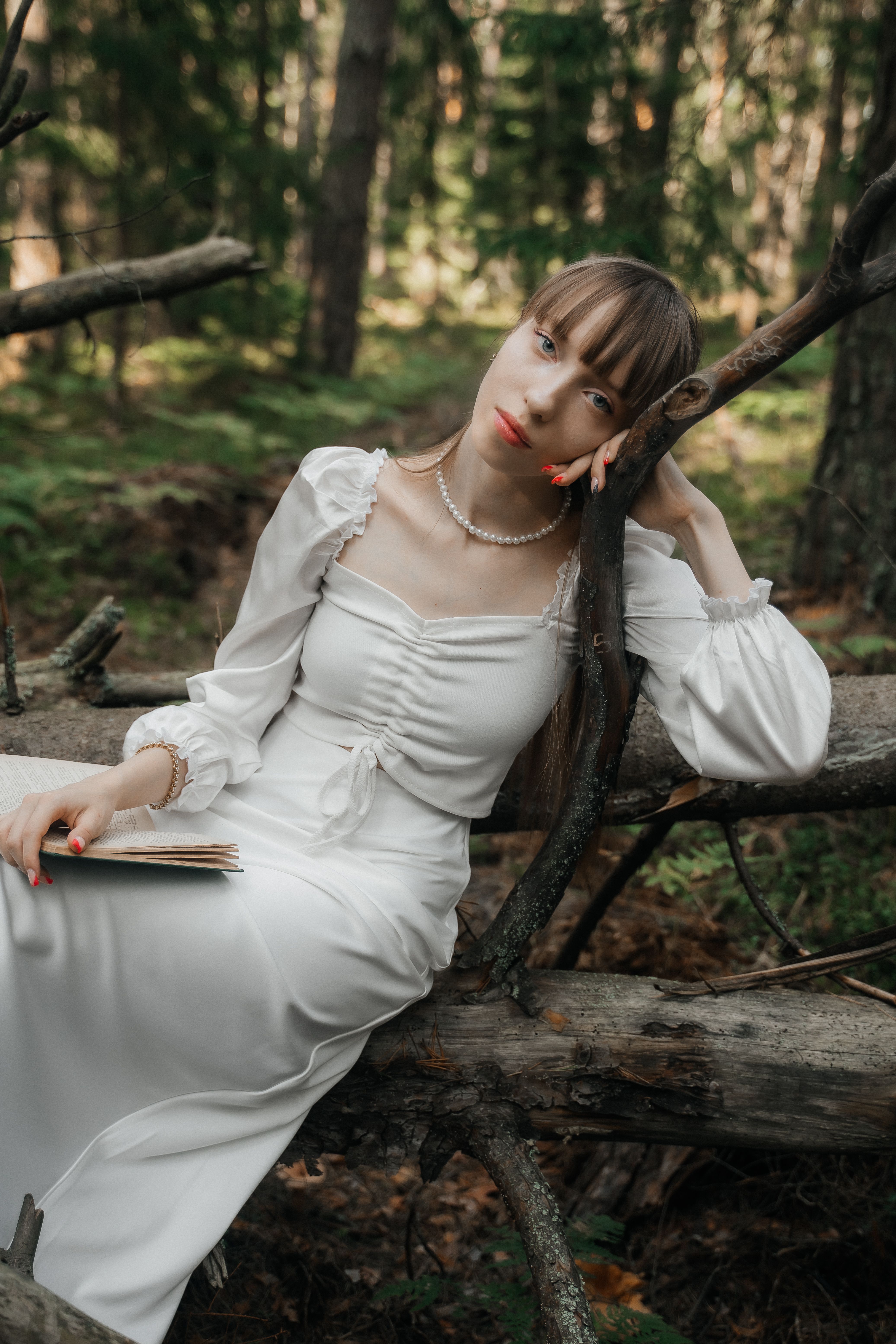 A young woman in a white dress rests on a fallen tree in a lush forest setting.