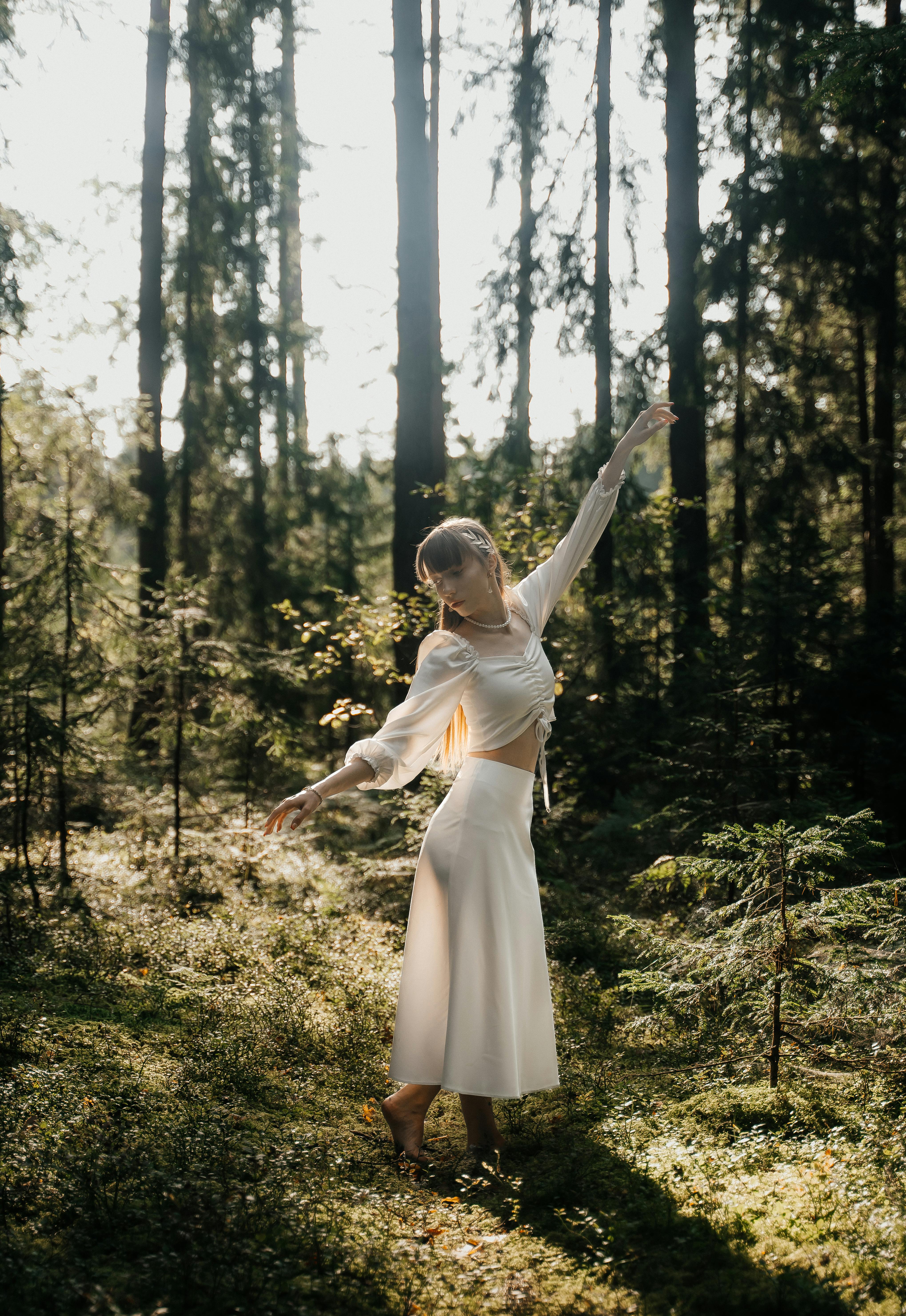 Serene woman in white dress gracefully dancing in a sunlit forest, embracing nature.