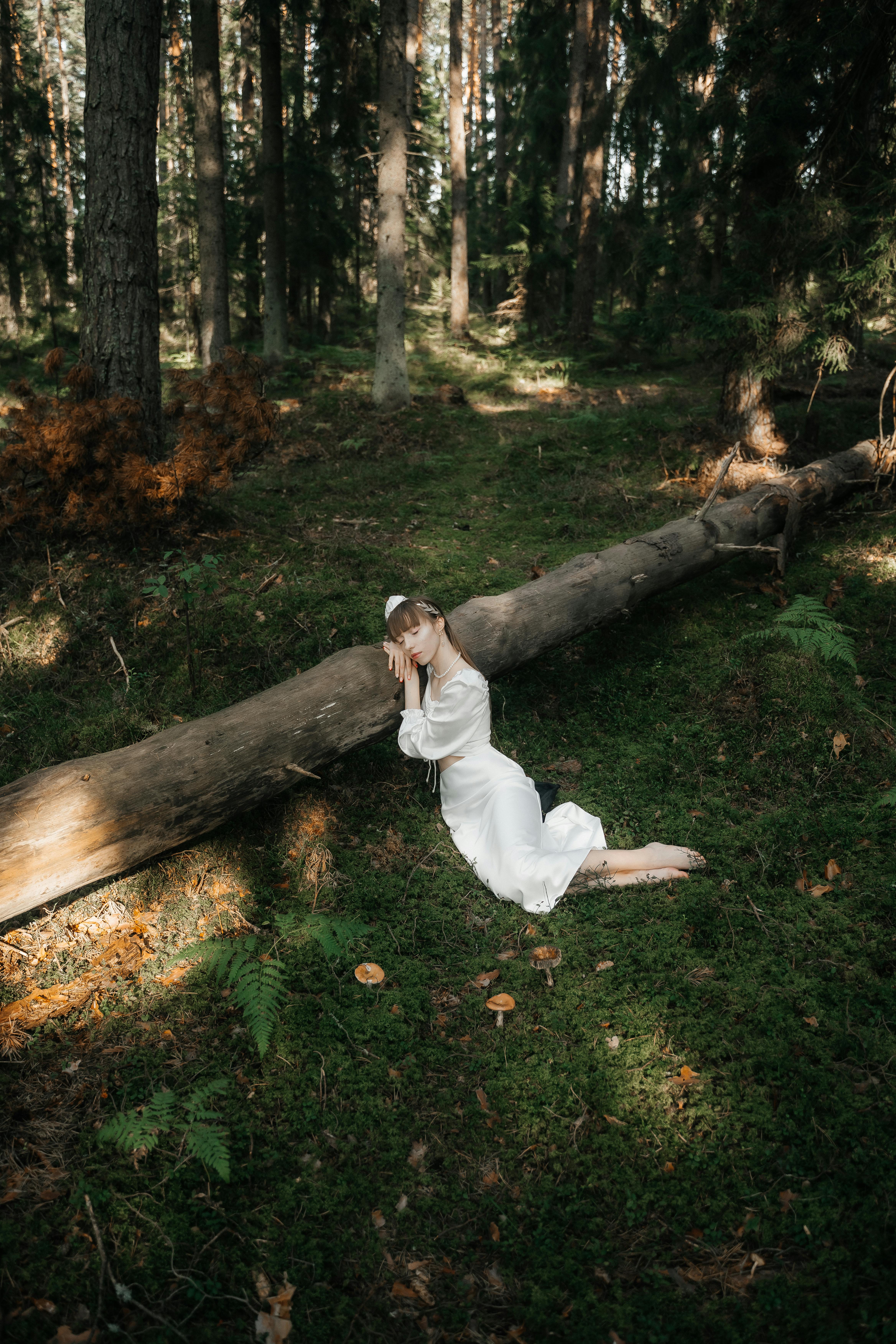Woman in white dress kneeling beside a fallen log in a serene forest.