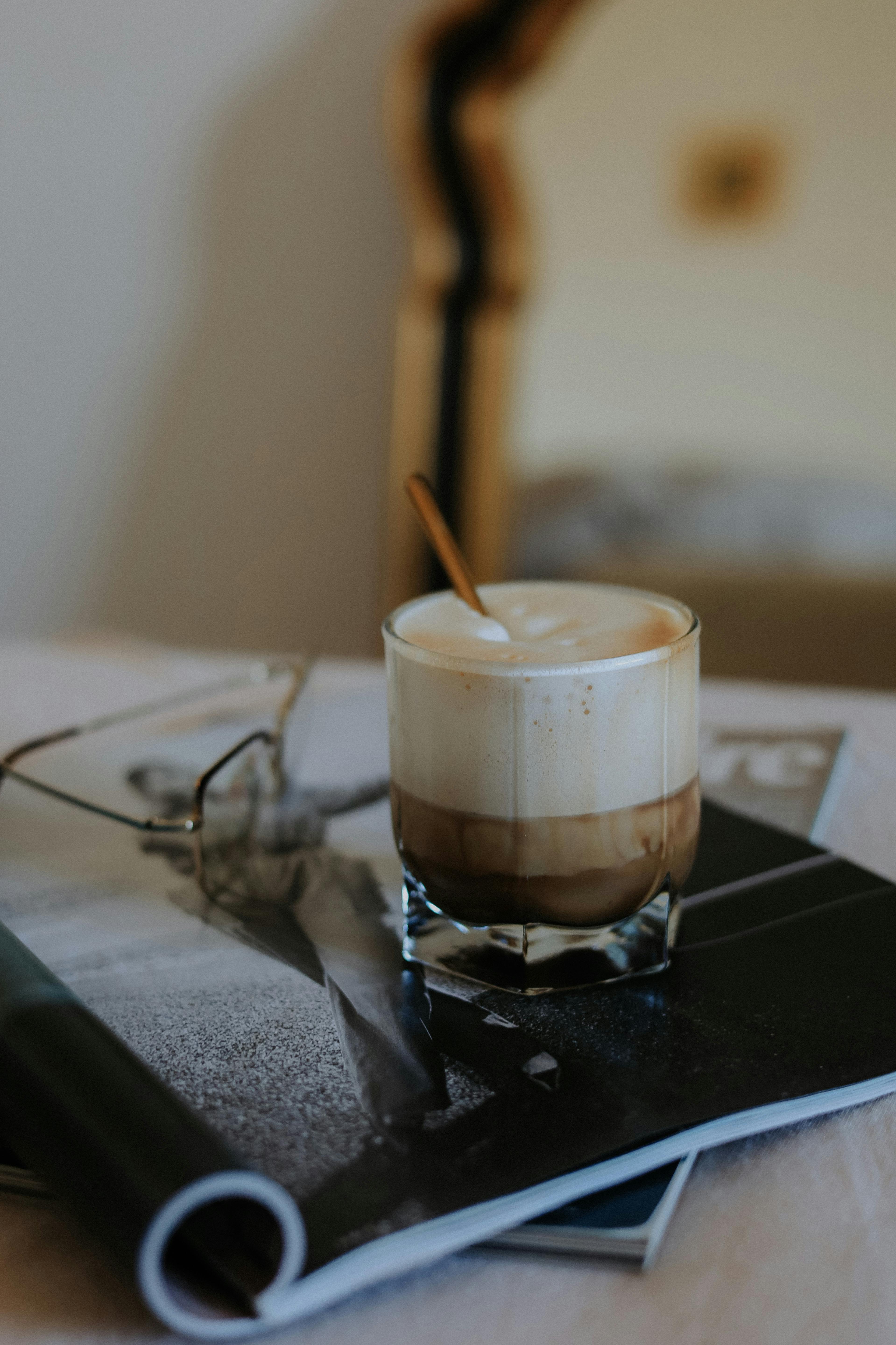 A comforting latte in a glass with spoon, surrounded by reading glasses and magazines. Ideal for cozy mornings.