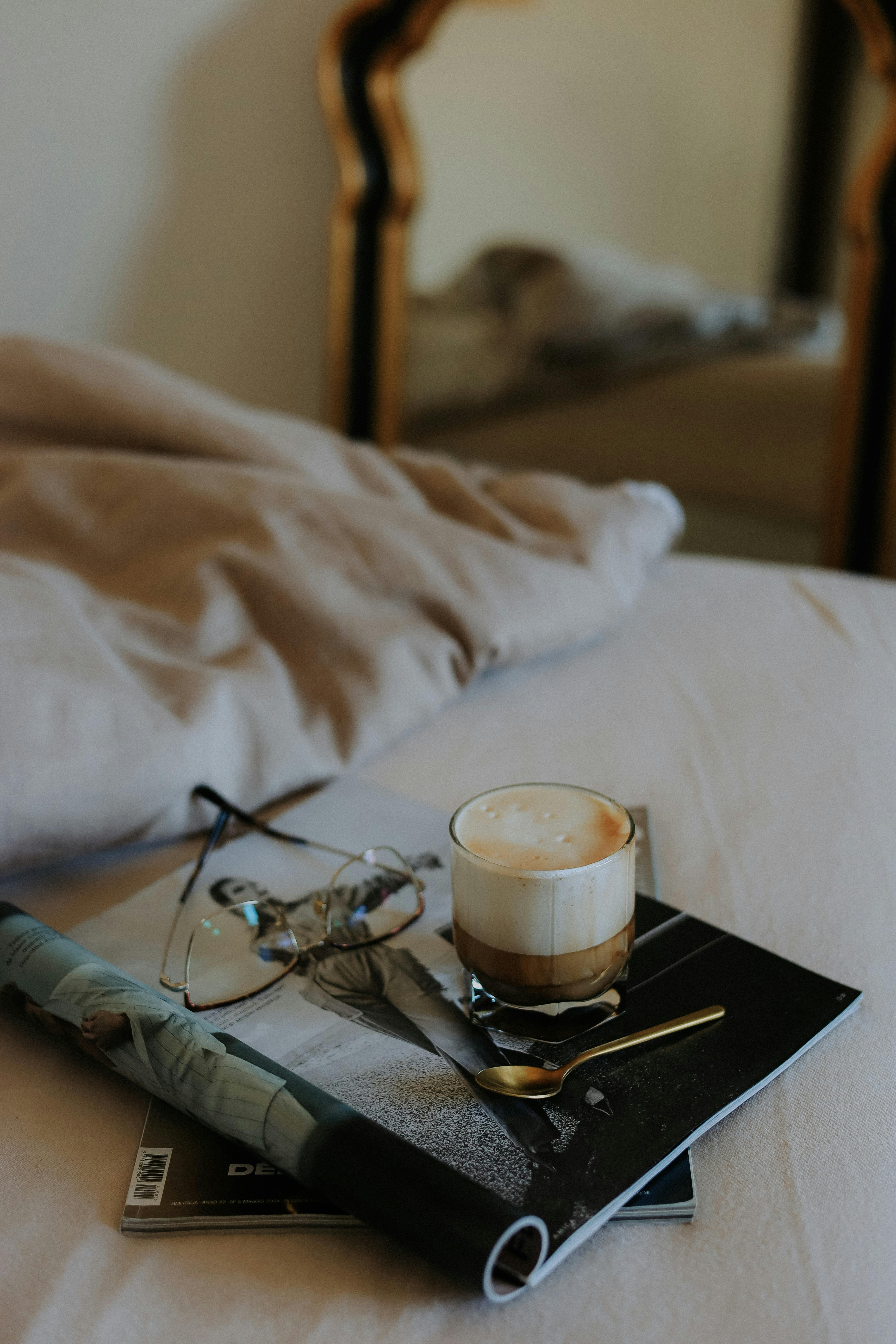 A latte with a spoon on a stack of magazines beside glasses, set on a cozy bed.