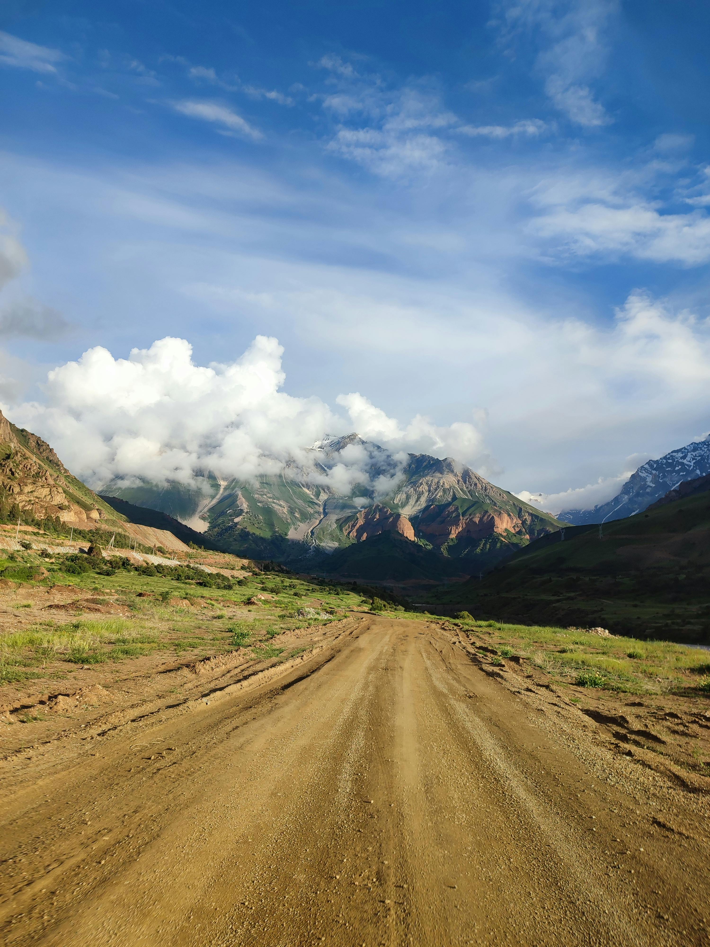A dirt road leading through a scenic mountain range with fluffy clouds and a blue sky.