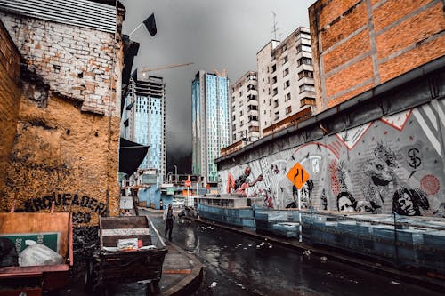 회색 하늘 아래 벽화가있는 건물을 따라 젖은 거리를 걷는 사람