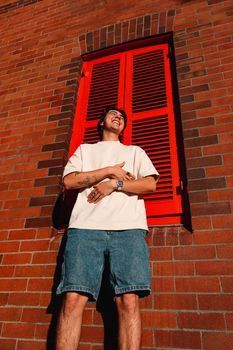 A young man in casual summer attire poses against a vibrant red-shuttered window in The Rocks, Sydney.