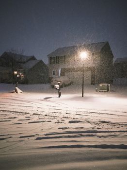 Snow falls at night in a quiet suburban neighborhood, creating a serene winter scene.