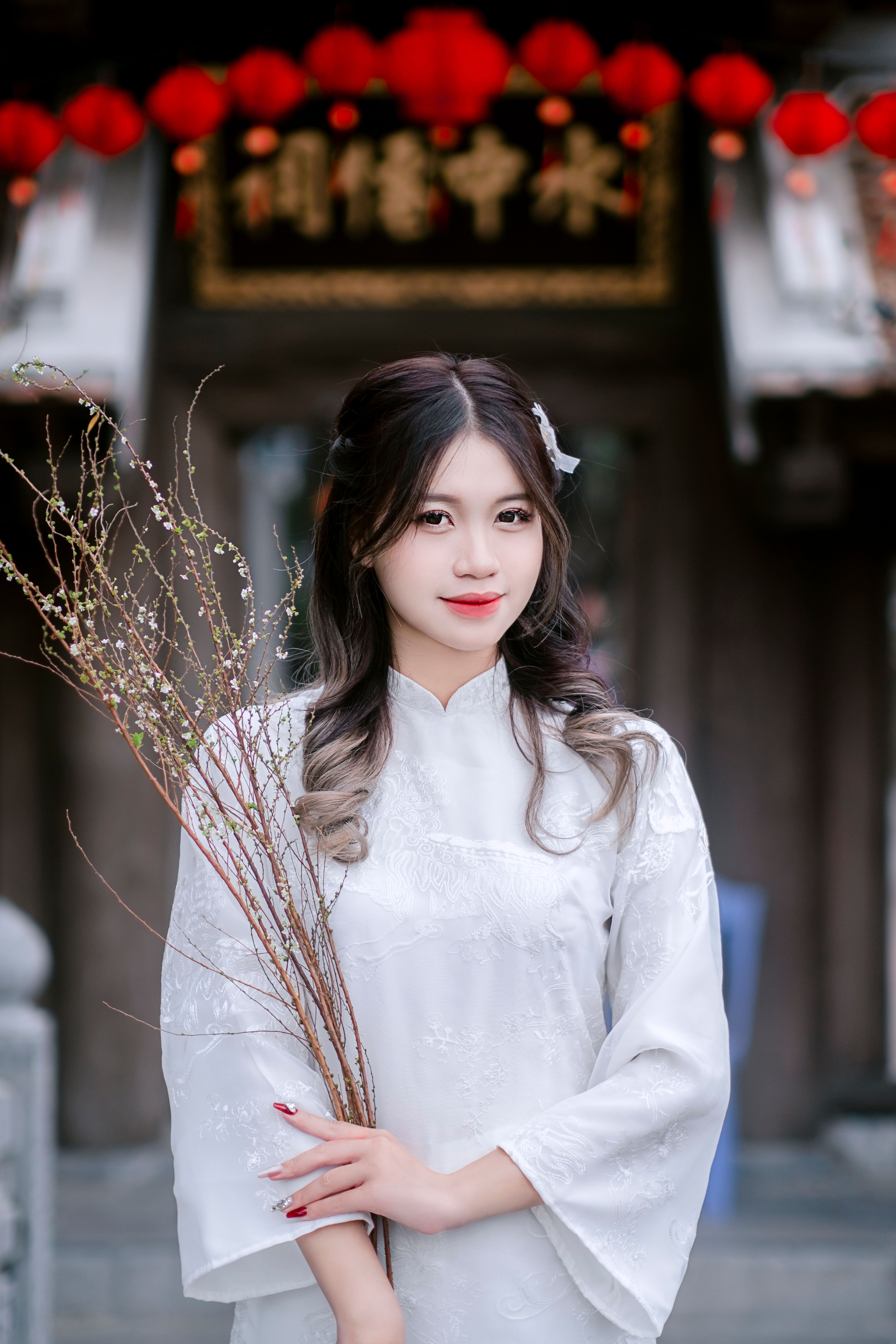 A young woman in traditional attire holding branches, standing in front of lanterns.