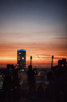 Silhouetted people admire a vibrant sunset with cityscape in the background, featuring a prominent skyscraper.