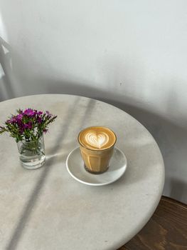 A latte with heart art and purple flowers on a minimalist round table.