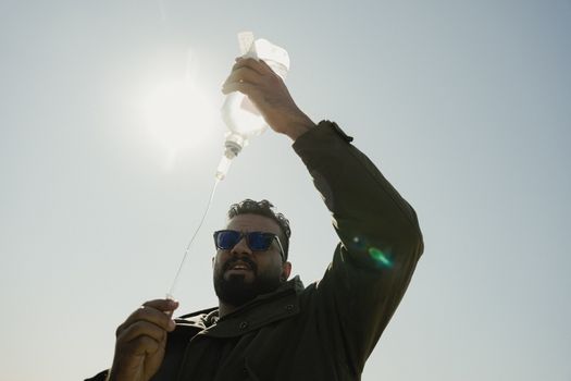 A man wearing sunglasses holds an IV bag outdoors under a clear sky, backlit by the sun.