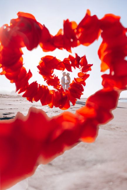 A couple embraces at the beach, framed by vibrant red petals forming a heart shape.