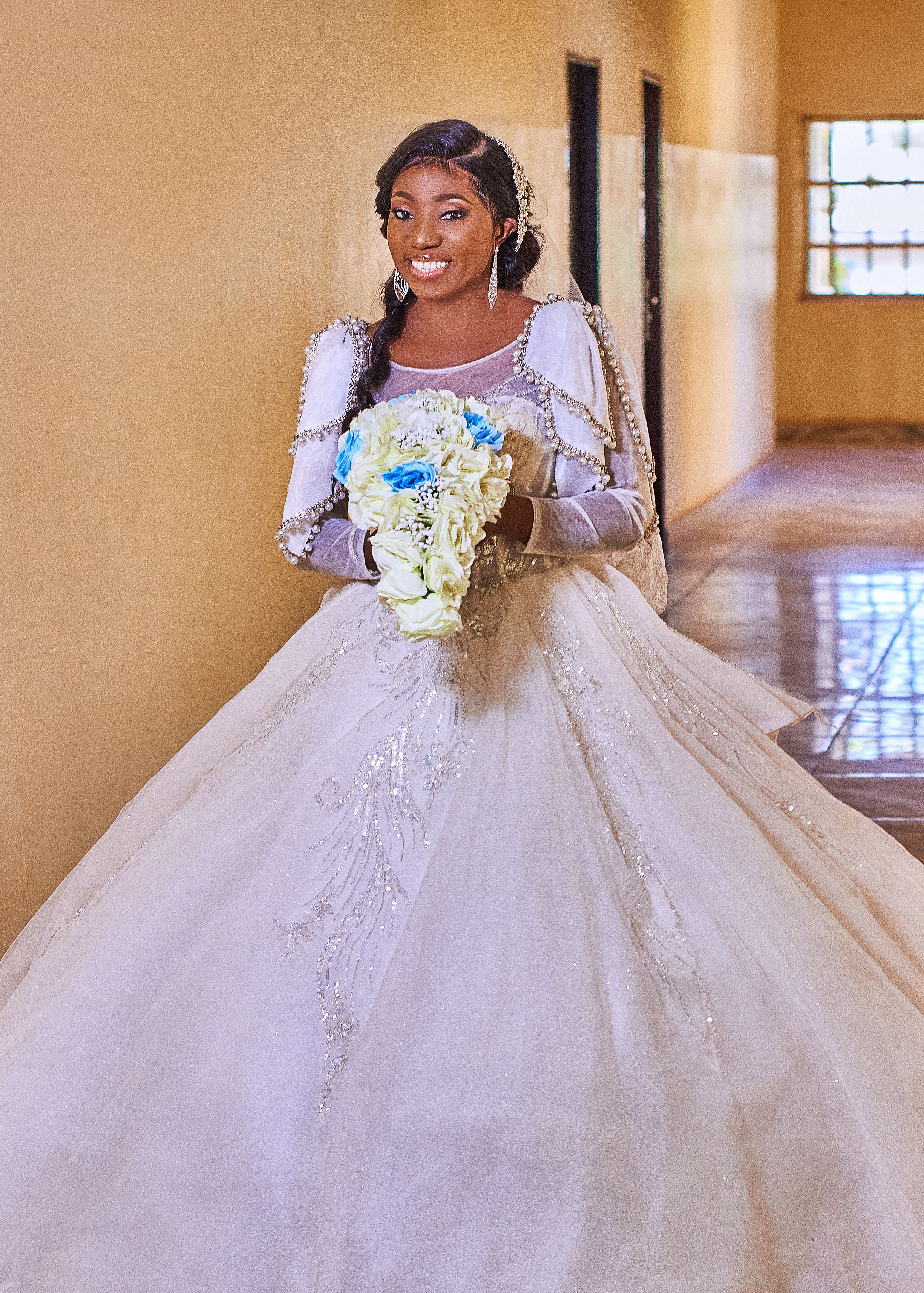 Smiling bride in white gown holding floral bouquet indoors.
