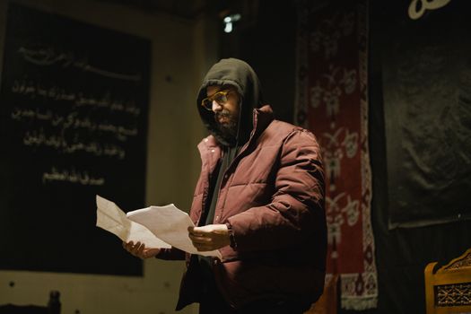 A man wearing a hooded jacket and glasses reads documents in a dimly lit room with decorative elements.