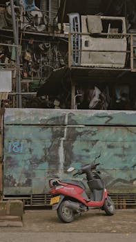 A red scooter parked near a scrapyard wall with industrial metal in the background.