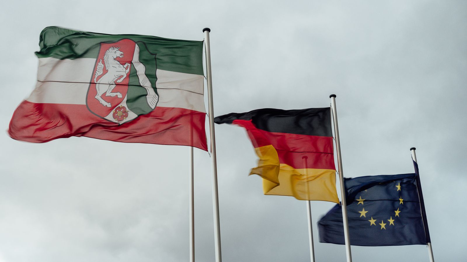 Flags of North Rhine-Westphalia, Germany, and the EU waving under a cloudy sky.