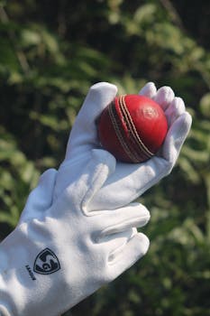 Close-up of a red cricket ball held in white gloves outdoors.