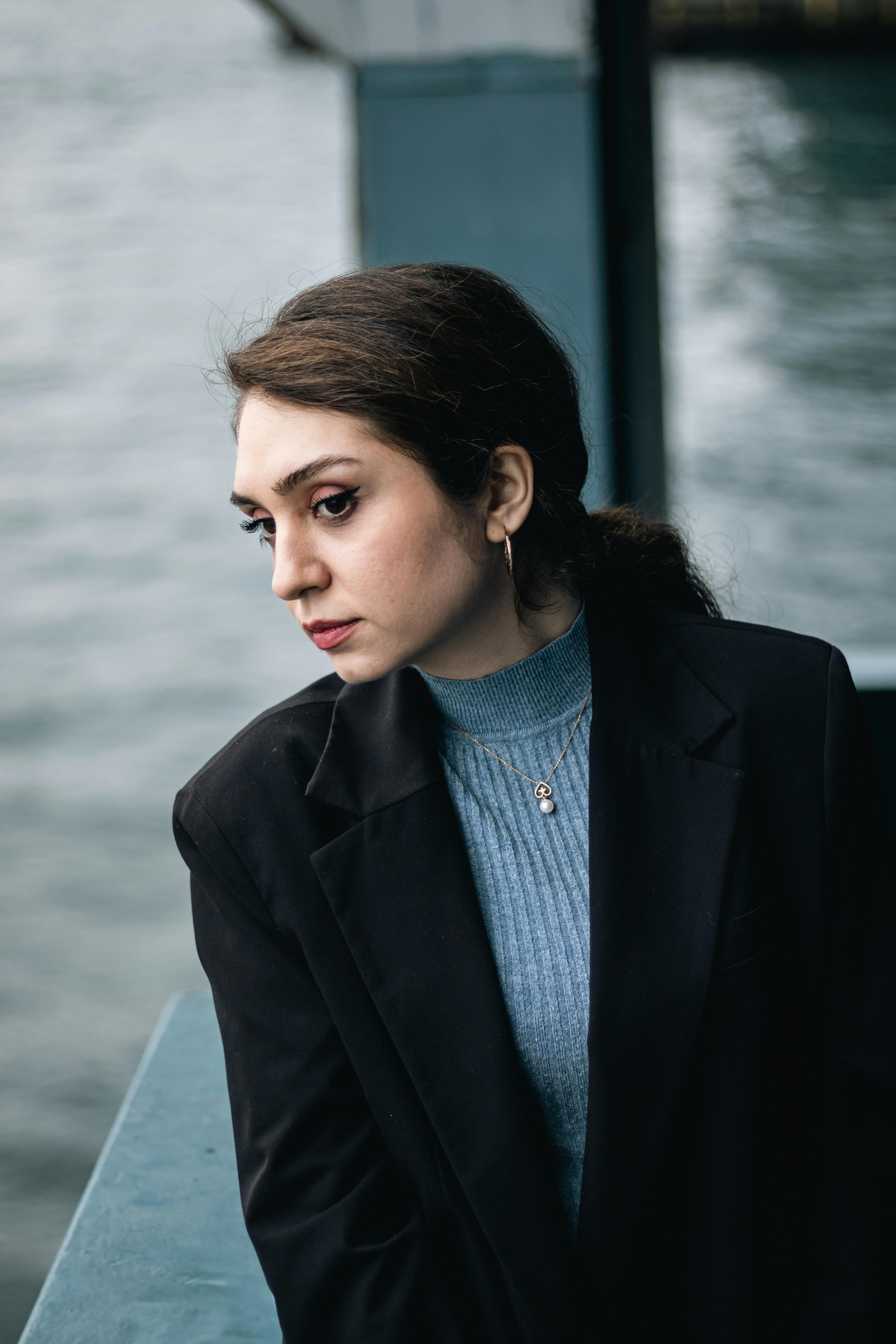 A thoughtful woman in a black blazer stands by the waterfront, gazing into the distance.