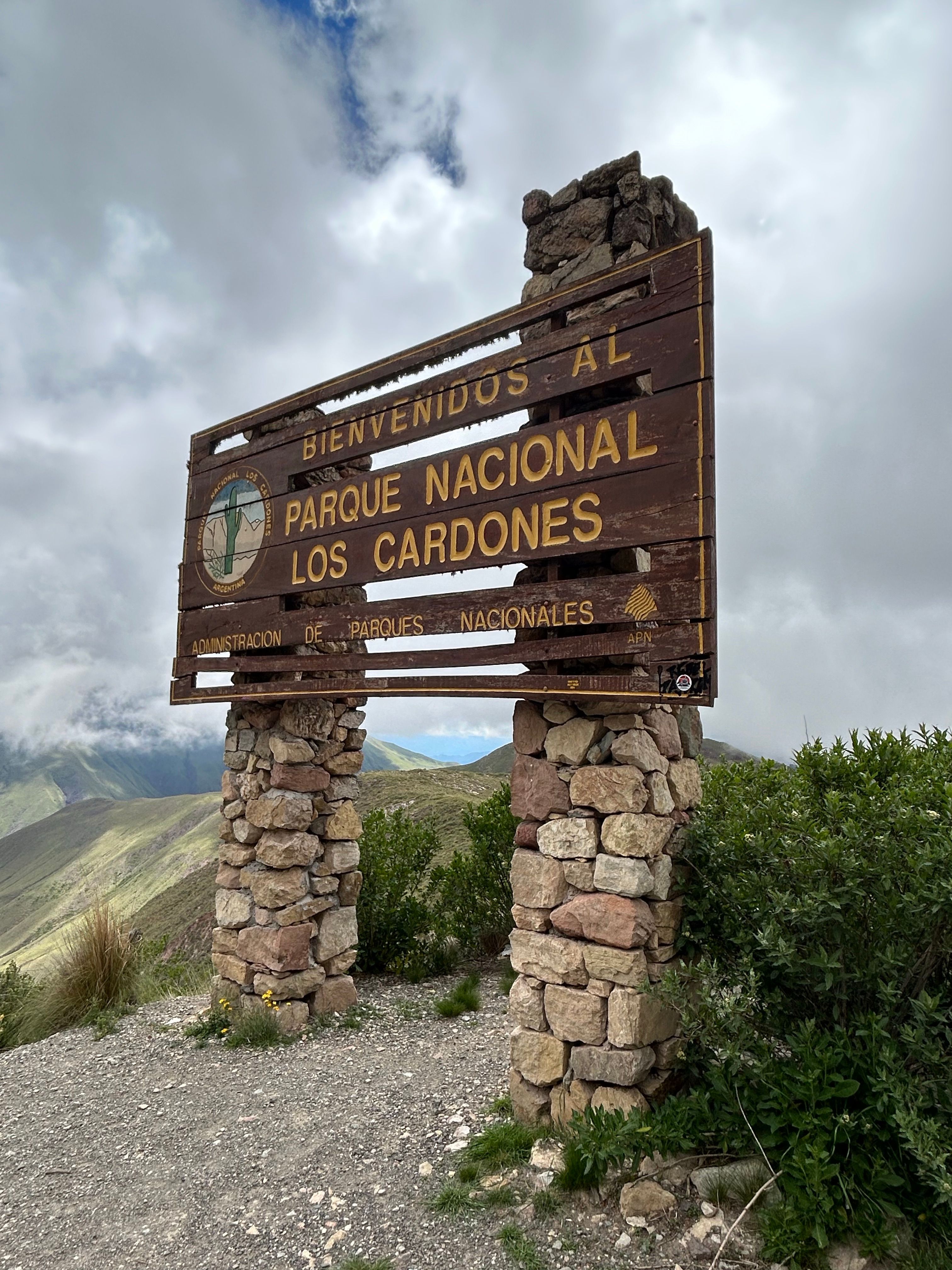 Fotomural Cartel de bienvenida en el Parque Nacional Los Cardones, Argentina, rodeado de frondosa vegetación y cielos nublados.