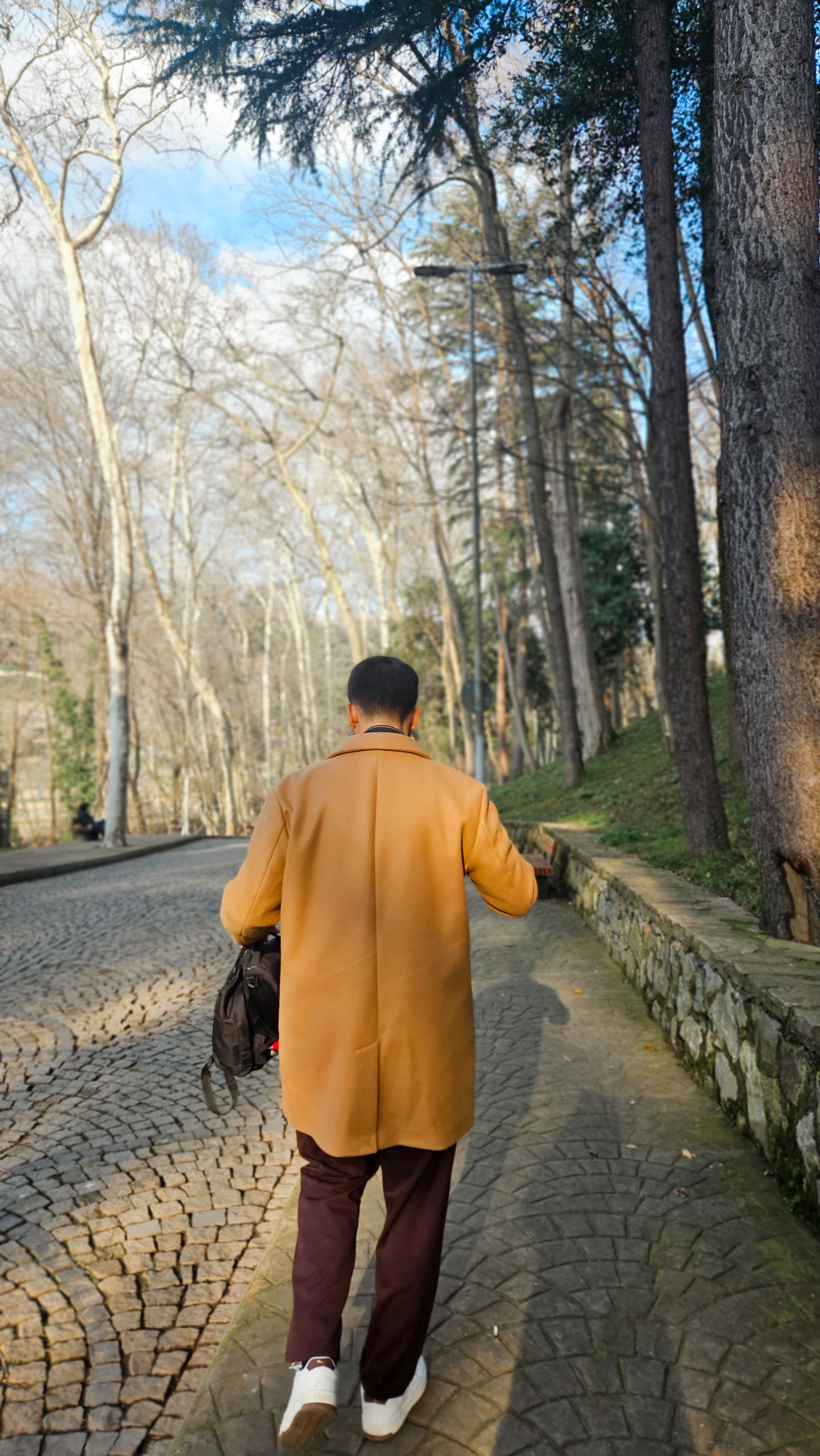 A man in a tan coat strolls on a cobblestone path surrounded by trees.