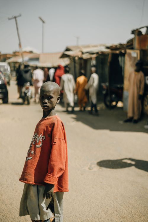 Free A young boy in a vibrant red shirt stands in a bustling street market. Stock Photo