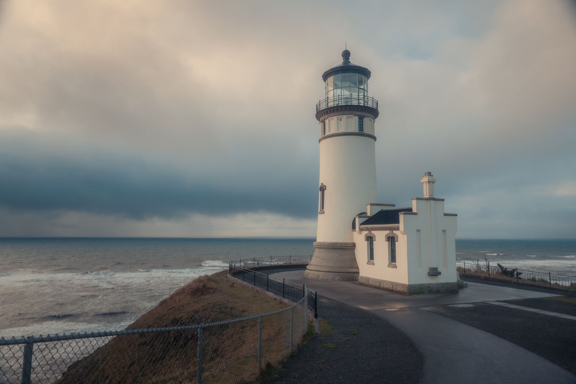 14 Lighthouses You Can Actually Spend the Night In 10 Peaceful scene of a historic lighthouse by the ocean under dramatic clouds.