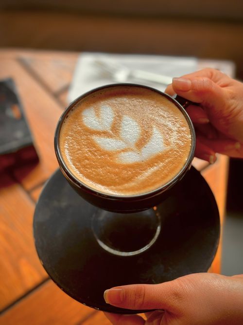 Free Close-up of latte art in a black cup, held by hands on a wooden table. Stock Photo