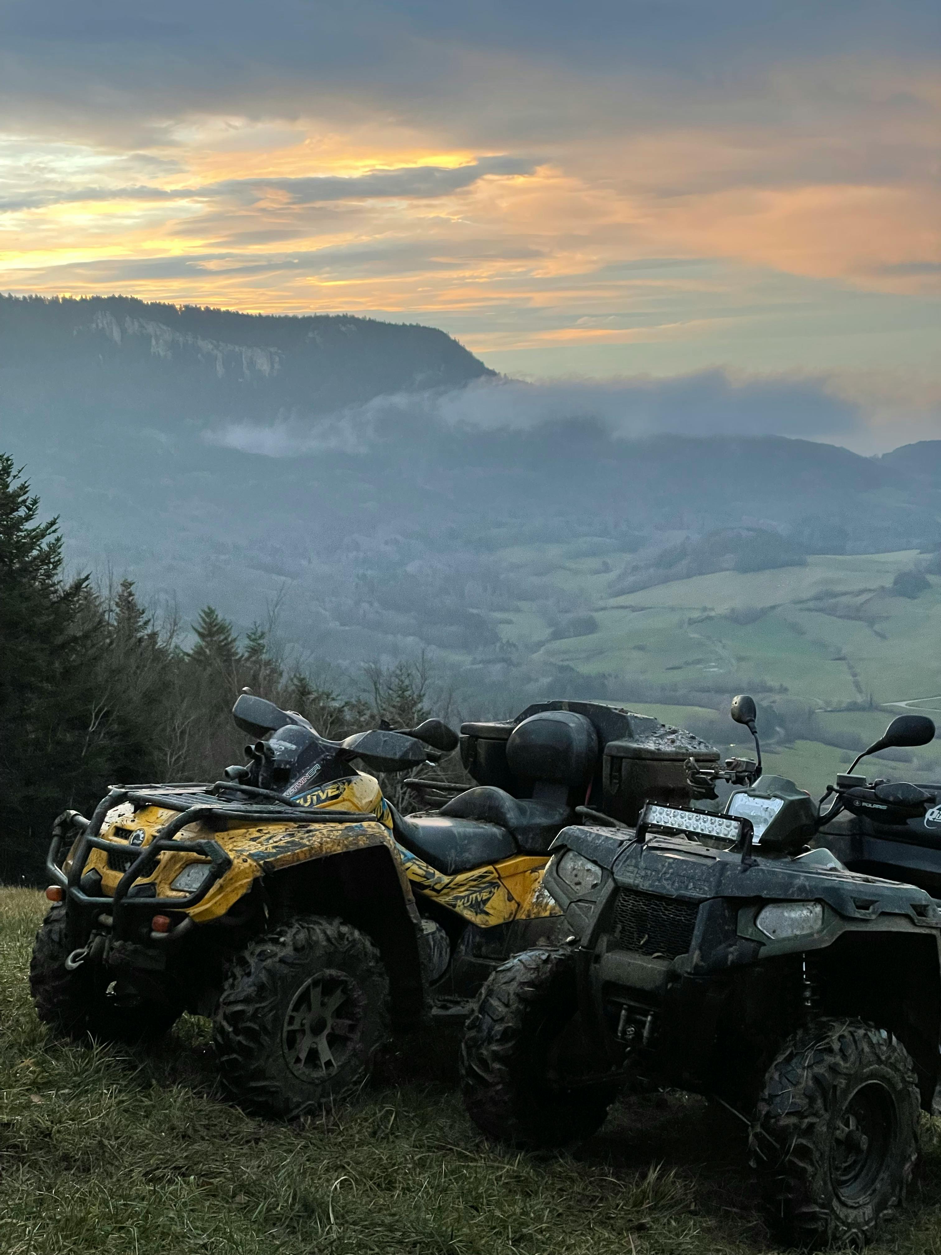 Two ATVs on a grassy mountain overlooking scenic misty valleys at sunset.