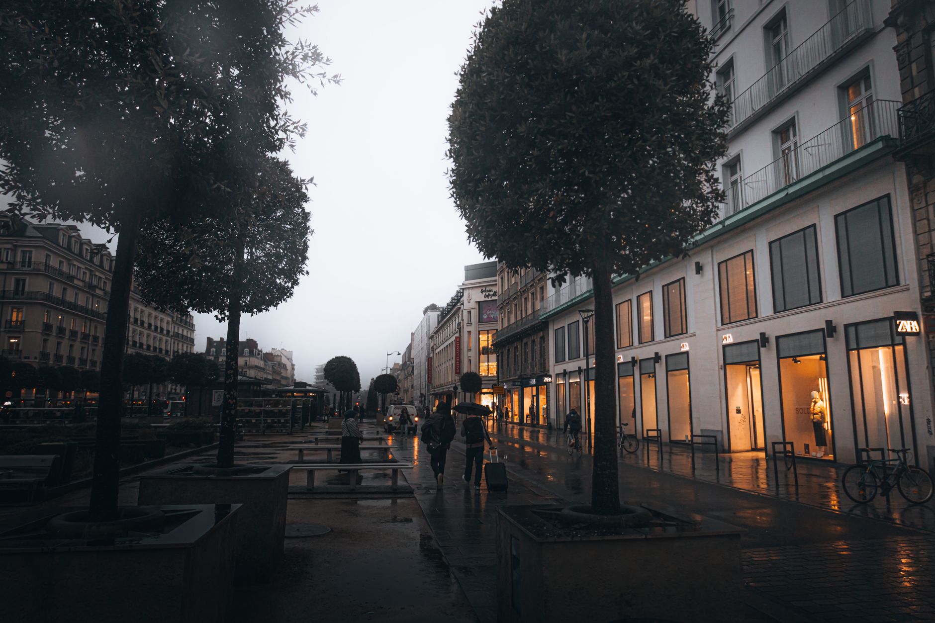People walking under umbrellas in a rainy, modern city street during twilight, reflecting city lights.