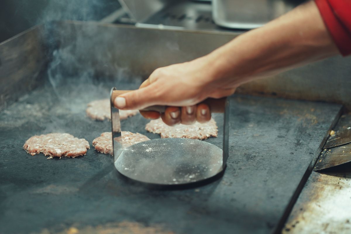 A cook pressing burger patties on a hot griddle.