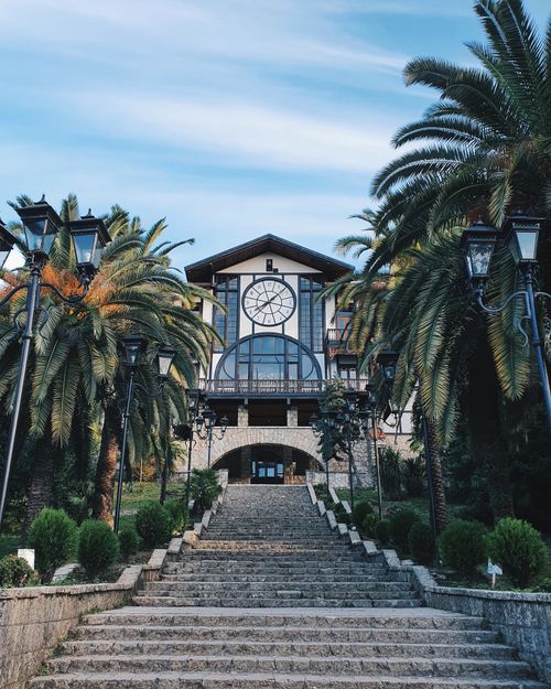 Free Elegant mansion entrance with clock, surrounded by palm trees, steps leading to it. Tranquil scenery. Stock Photo