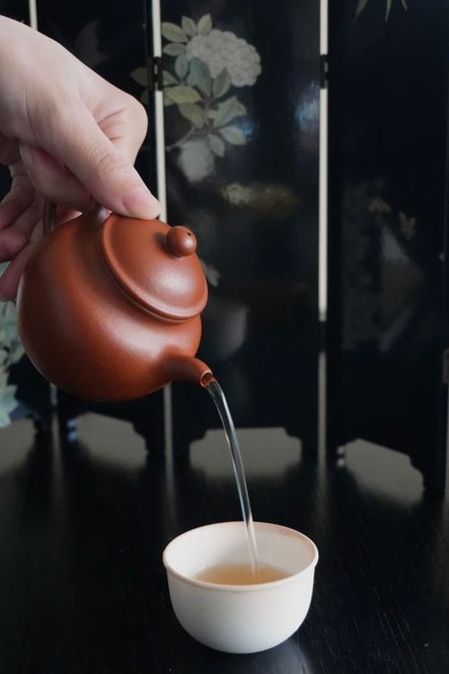 Free A hand pours tea from a ceramic teapot into a small teacup, indoors. Stock Photo