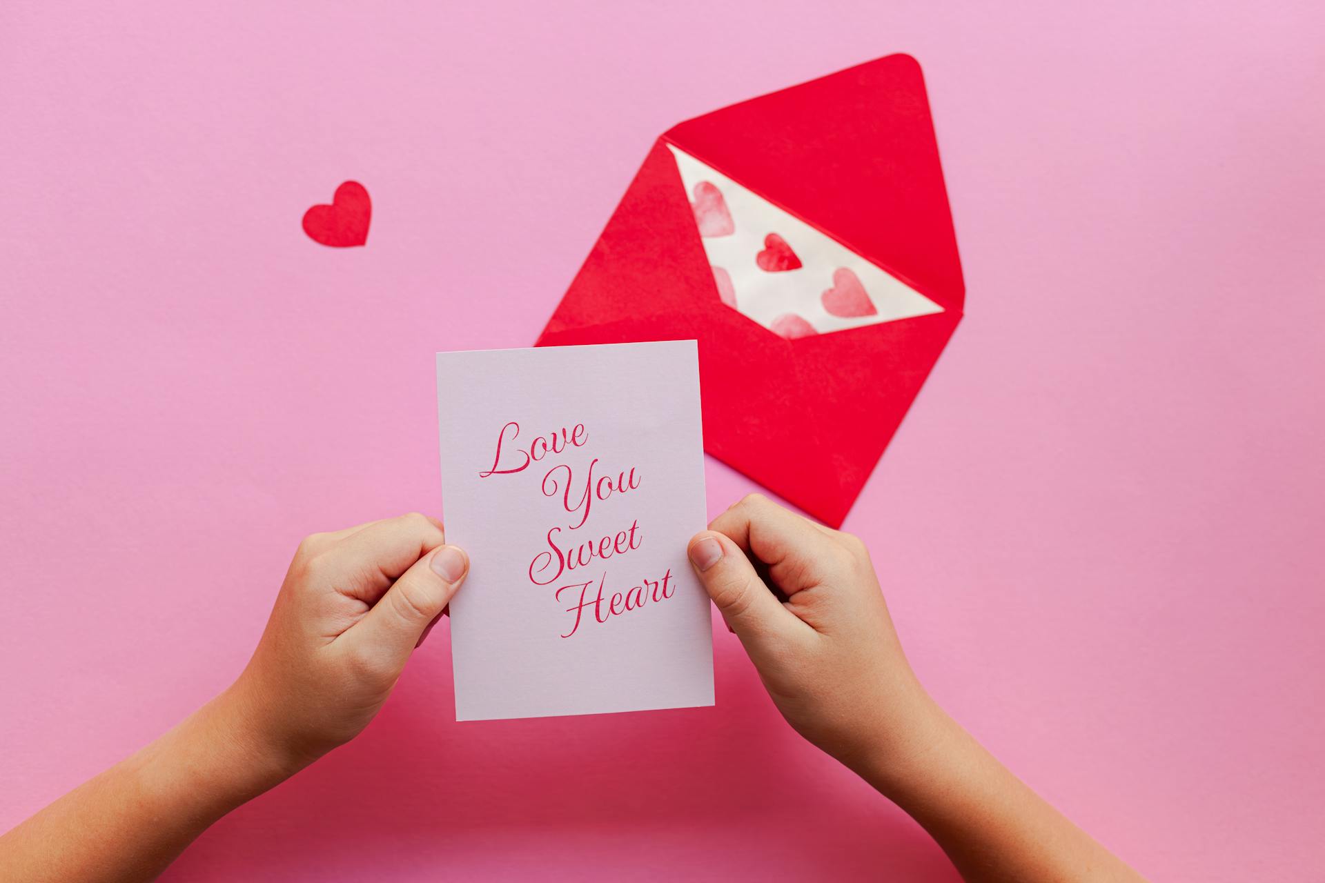 Hands holding a romantic greeting card above a pink background with a red envelope.