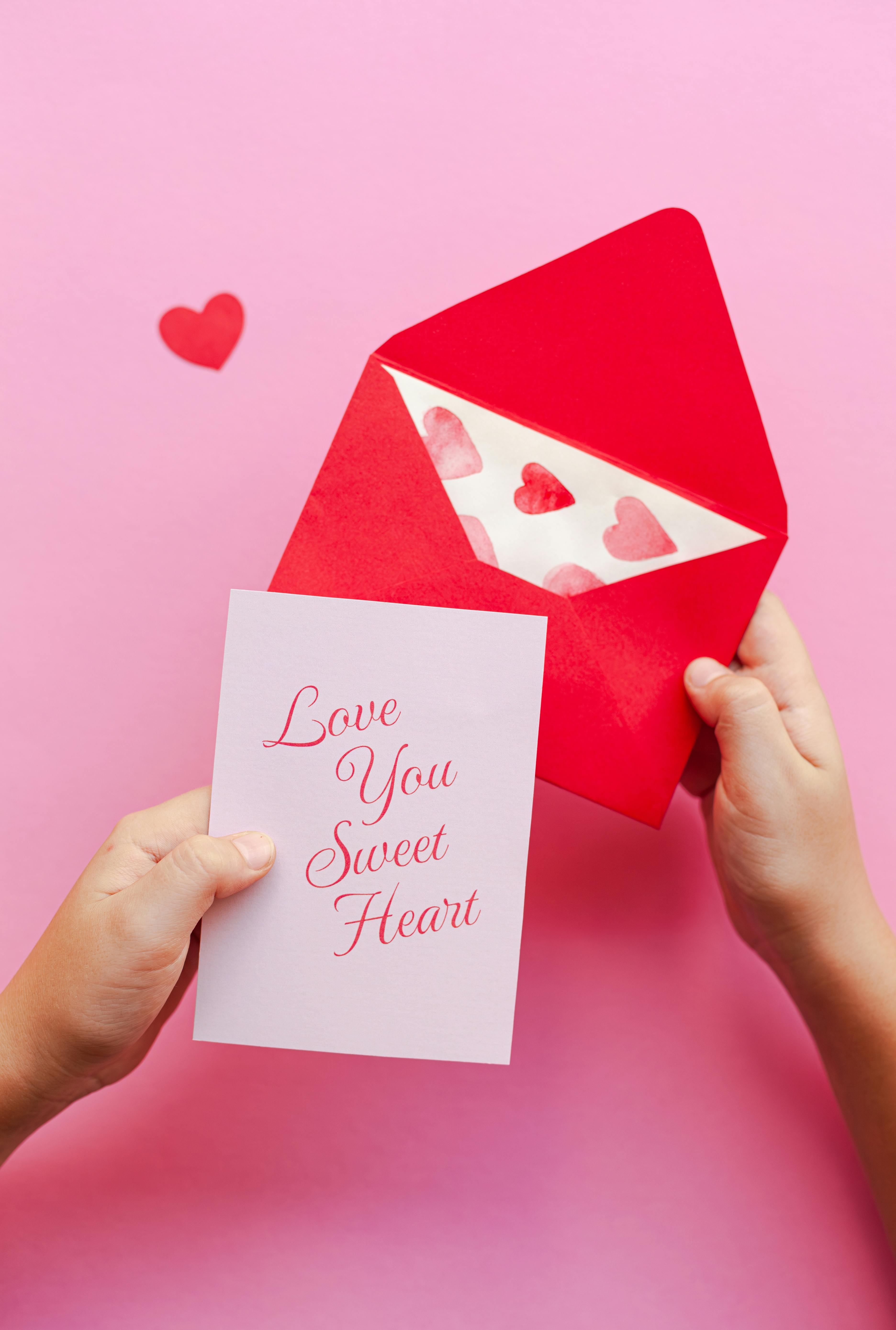 Hands hold a love-themed card and red envelope on a pink background.