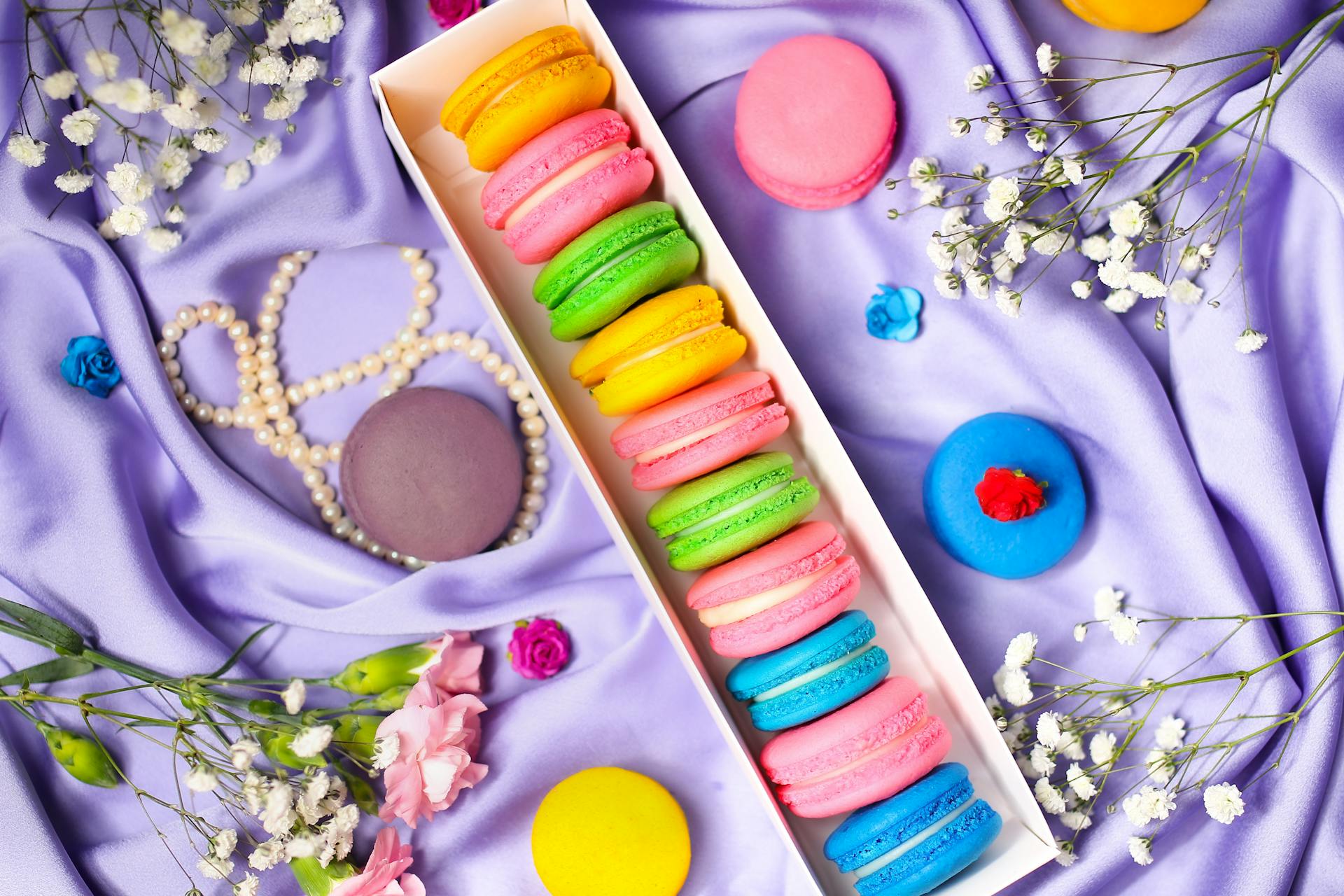 Vibrant macarons arranged on lilac fabric with delicate flowers and pearls.