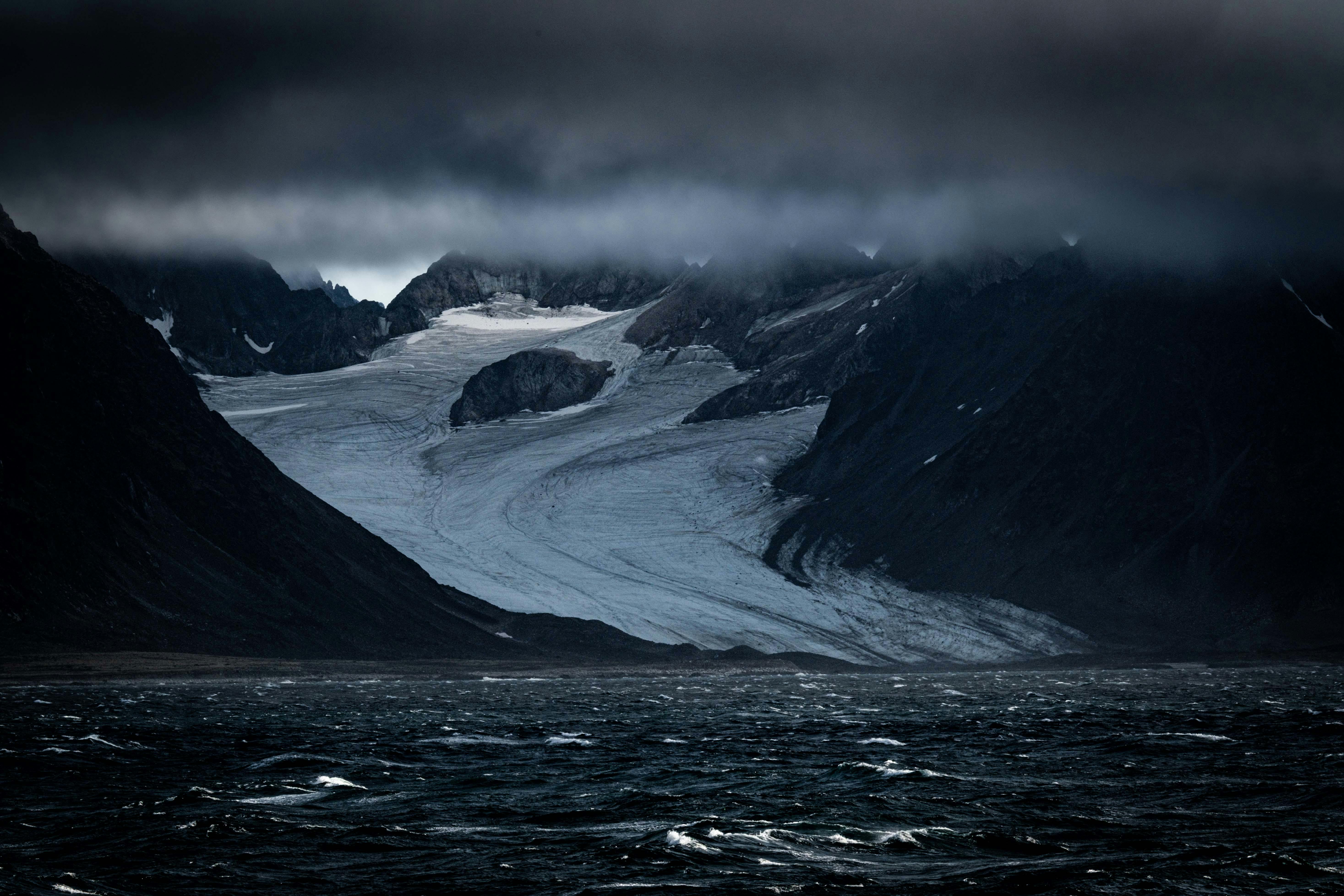 A breathtaking view of a glacier flanked by dark mountains beneath dramatic clouds over turbulent waters.
