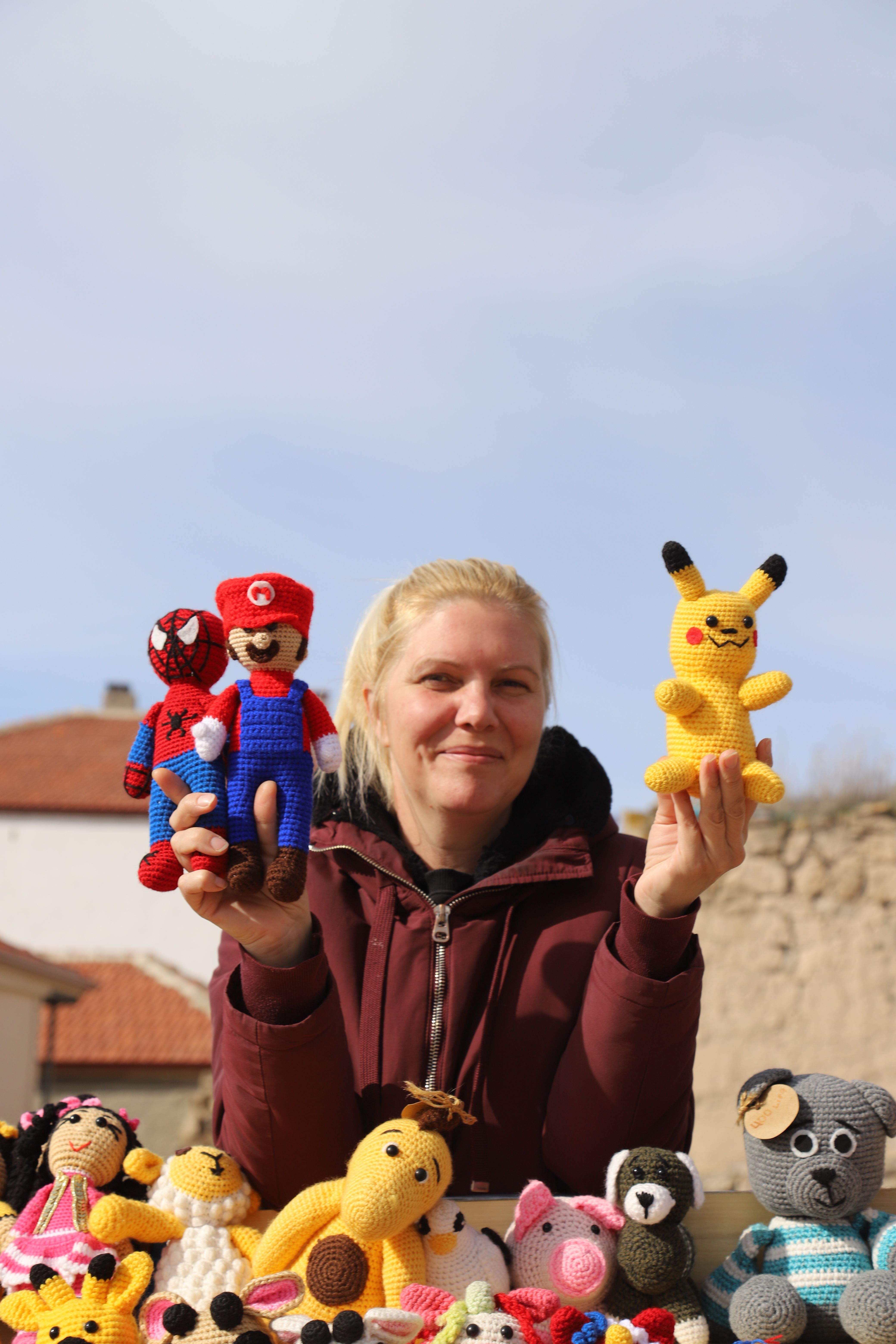 Woman showcasing colorful crochet toys including Pikachu under blue sky.