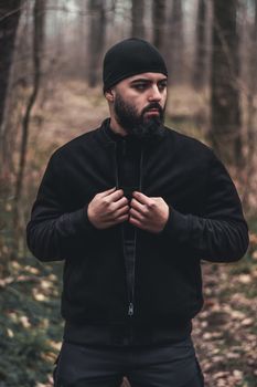 A bearded man dressed in a black jacket stands in an autumn forest, exuding a moody and rugged vibe.