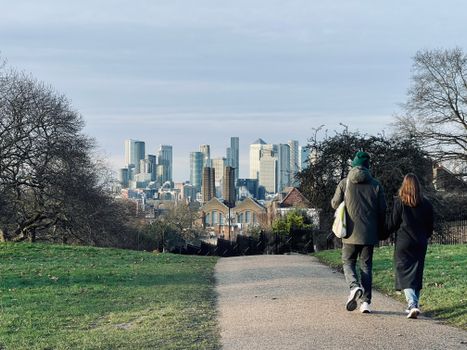A couple walks through Greenwich Park overlooking London's skyline on a clear day.