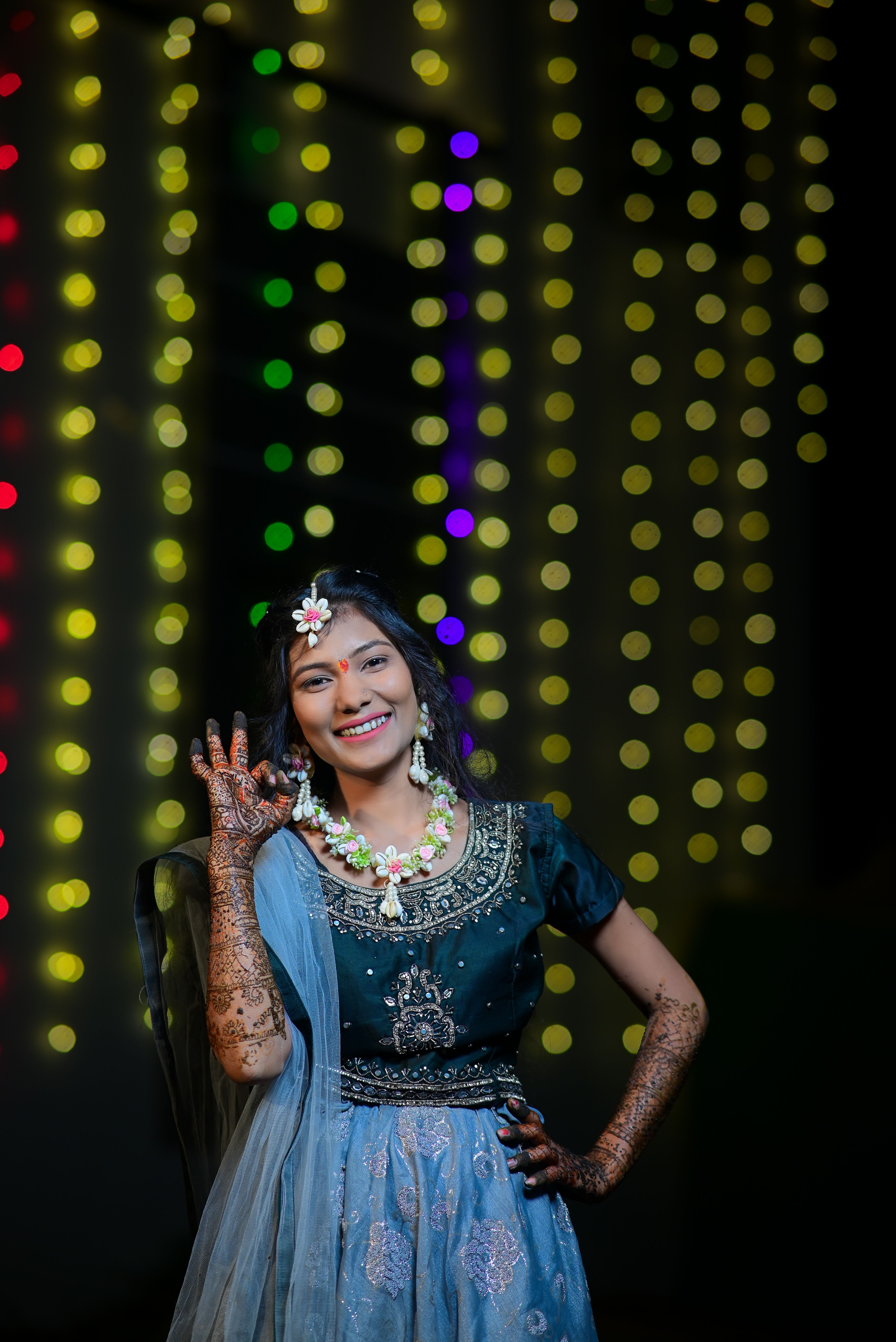 Smiling woman in traditional Indian attire adorned with henna and floral jewelry against festive lights.