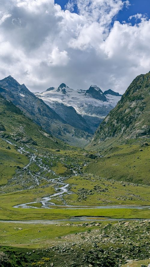 Free A picturesque alpine valley with a meandering stream and snow-capped mountains under a cloudy sky. Stock Photo