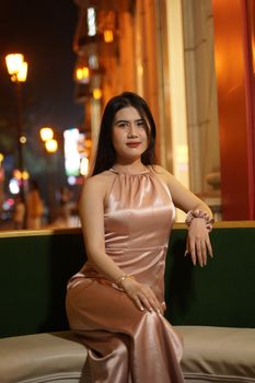 A woman in a satin dress posing elegantly at night with city lights in the background.