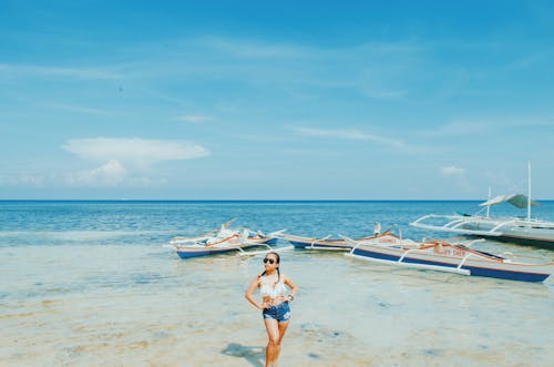 Женщина в белом купальнике и синих коротких шортах стоит под акимбо обеими руками у моря под синим и белым небом