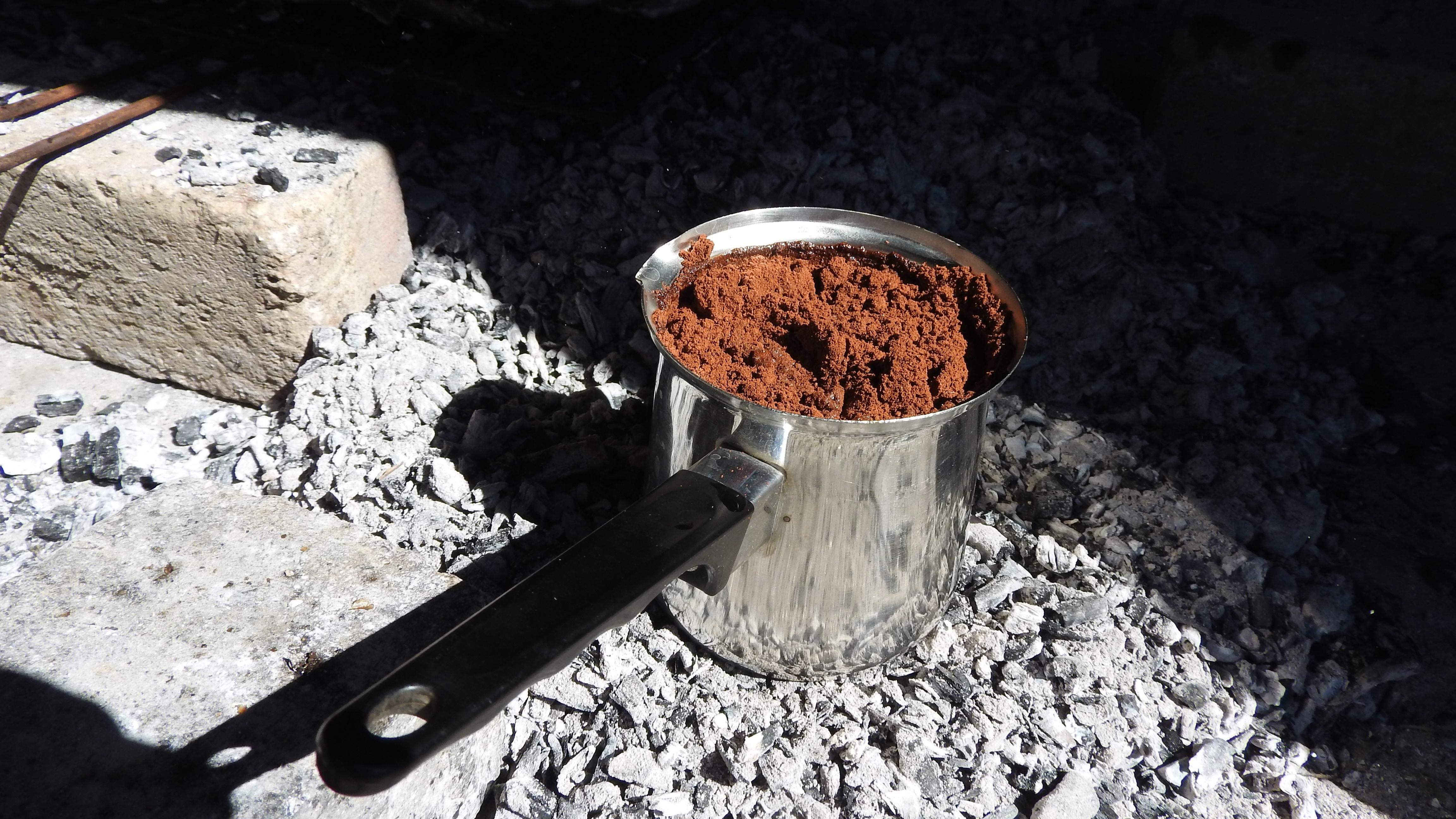 Metal pot filled with ground coffee placed on charcoal outdoors, in bright sunlight.