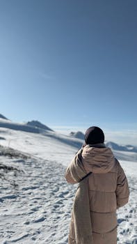 Person bundled in winter attire hiking on a snowy mountain trail under a clear sky.