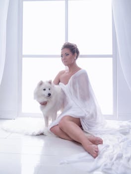 A woman in white drapery poses with a fluffy dog by a window, creating a serene ambiance.