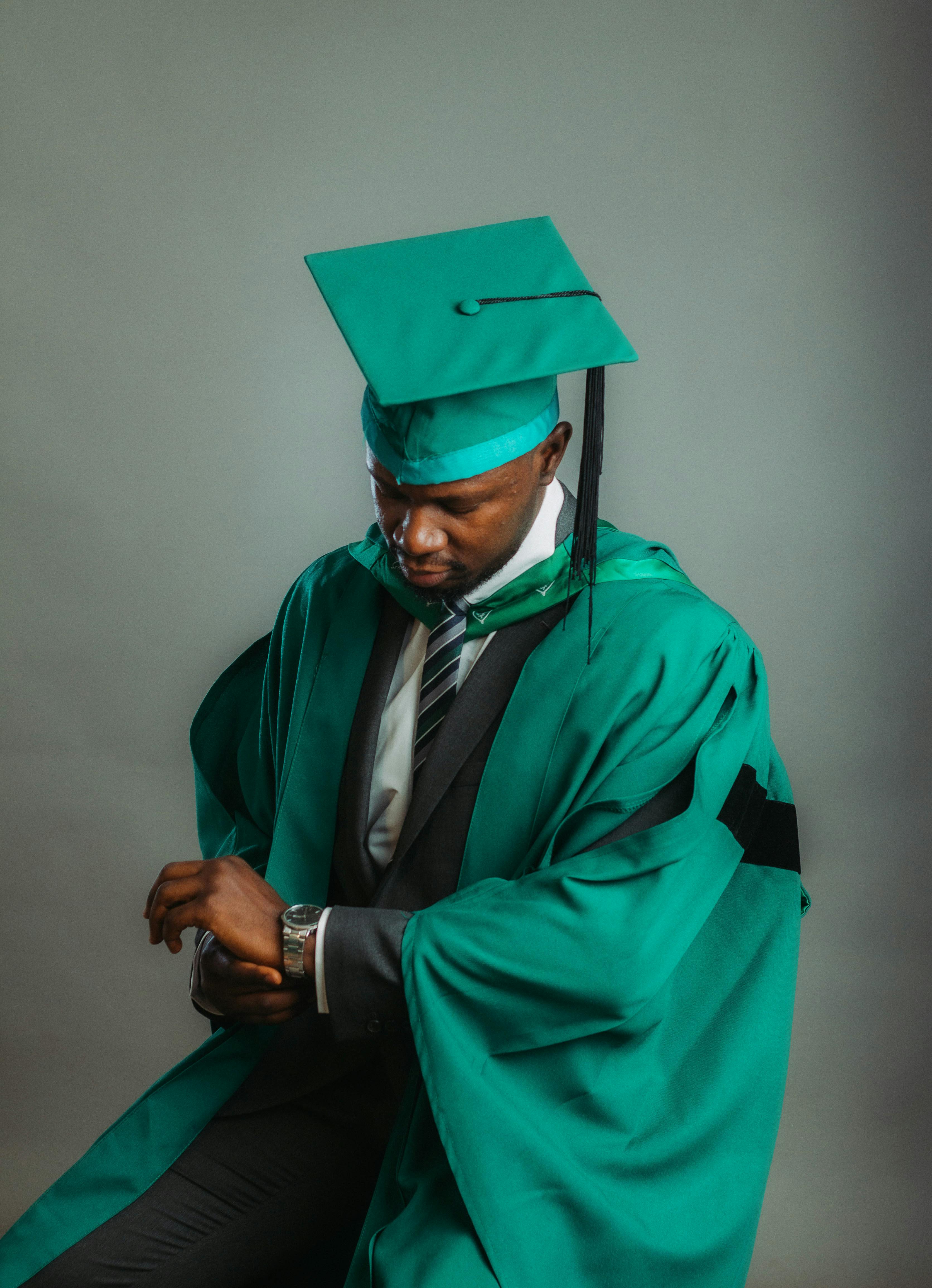Portrait of a young African graduate wearing a green cap and gown, looking at watch.