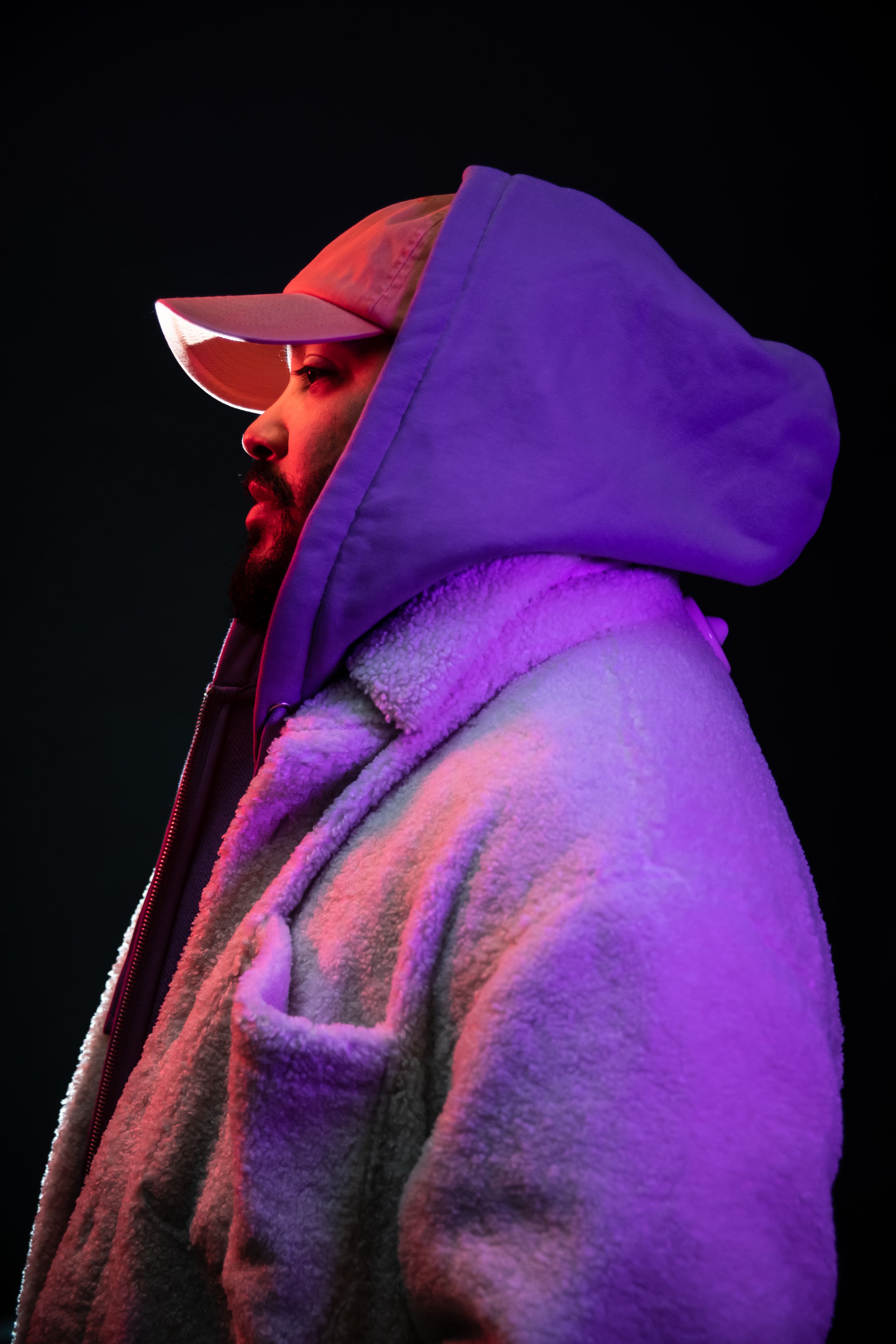 Side profile of a bearded man wearing a hooded jacket under vibrant neon lights.